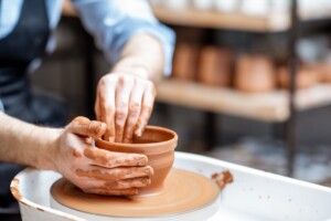 Man making clay jug forming shape by hands on the pottery wheel indoors, close-up view
